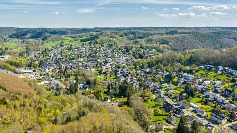 A picturesque view of a peaceful village surrounded by green hills and forests. The houses are well distributed, and the landscape appears inviting and serene.
