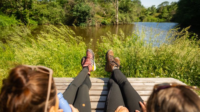Twee mensen ontspannen op een bankje aan de oever van een vijver, omgeven door groene natuur.
