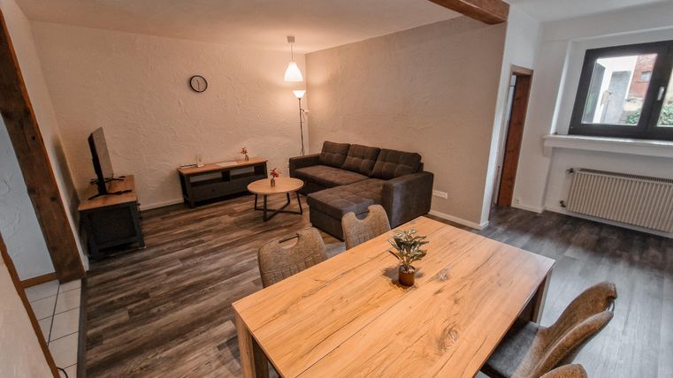 Ein modernes Wohnzimmer mit einem Sofa, einem Tisch und einer Lampe. Der Raum hat große Fenster und einen gemütlichen Holzfußboden.
