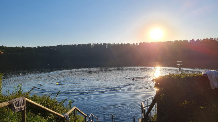 A tranquil lake with gentle waves beneath a clear sky. The sun rises on the horizon, illuminating the surroundings.