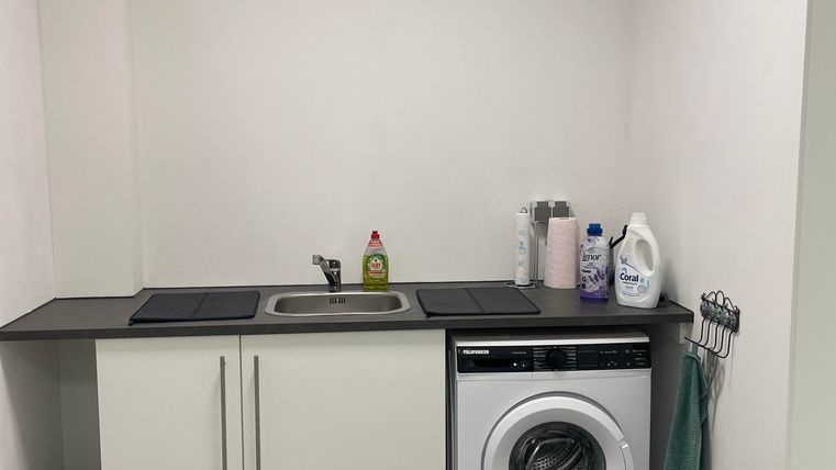 Ein helles Waschzimmer mit einer Waschmaschine und einem kleinen Waschbecken. An der Wand hängen Reinigungsmittel und Handtücher.