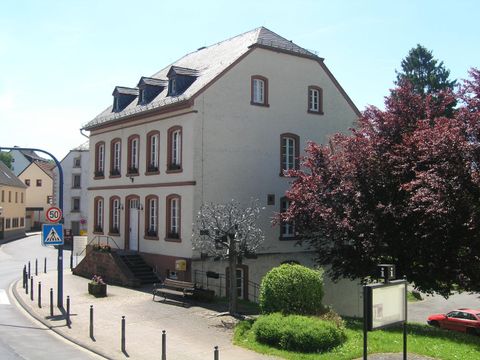 A two-story, bright building with white walls and a sloping roof. In the foreground, there is a green lawn and some trees.