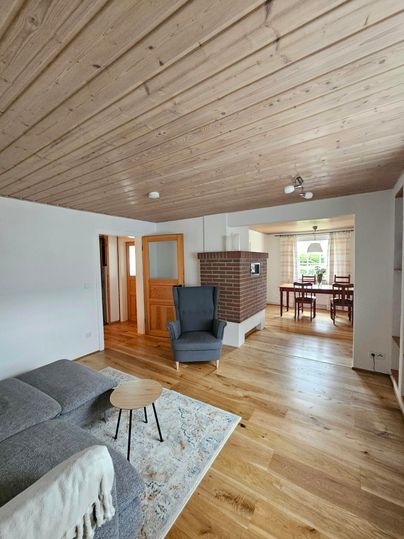 Een lichte woonkamer met houten vloeren en moderne meubels. Op de achtergrond is een eettafel zichtbaar.