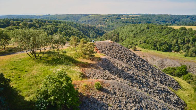 Blick auf drei Schieferhügel. Im Hintergrund sind grüne Wälder zu erkennen.