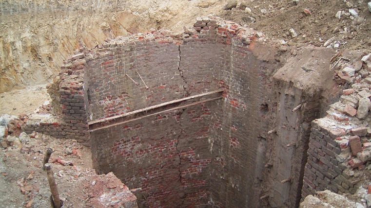 A deep shaft made of bricks that leads into the ground. The surrounding ground is uneven and shows earthworks.