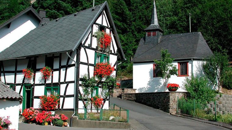 A picturesque village with traditional half-timbered houses and a small church. Colorful flowers adorn the buildings, and the surroundings are green and inviting.