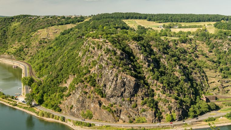 Eine malerische Landschaft mit sanften Hügeln und bewaldeten Gebieten. Im Vordergrund verläuft ein Fluss entlang der Uferstraße.