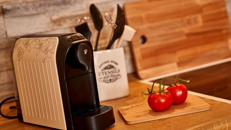 Eine moderne Kaffeemaschine steht auf einem Holztisch. Neben ihr liegen frische Tomaten und Küchenutensilien.