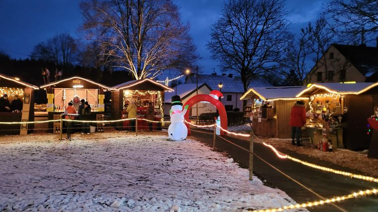 Ein Weihnachtsmarkt bei Nacht mit festlich beleuchteten Ständen und einem Schneemann. Der Boden ist mit Schnee bedeckt und es sind Menschen zu sehen, die den Markt besuchen.