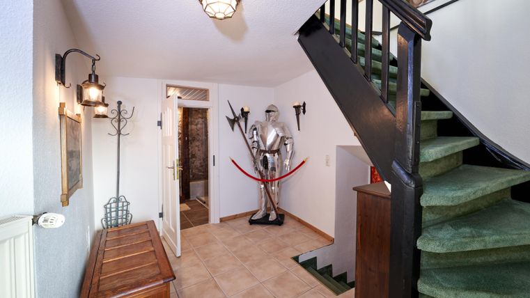 Ein Eingang mit einer Treppe und einem Holzschrank. Im Mittelpunkt steht eine Rüstung, umgeben von alten Möbeln und einer gemütlichen Atmosphäre.
