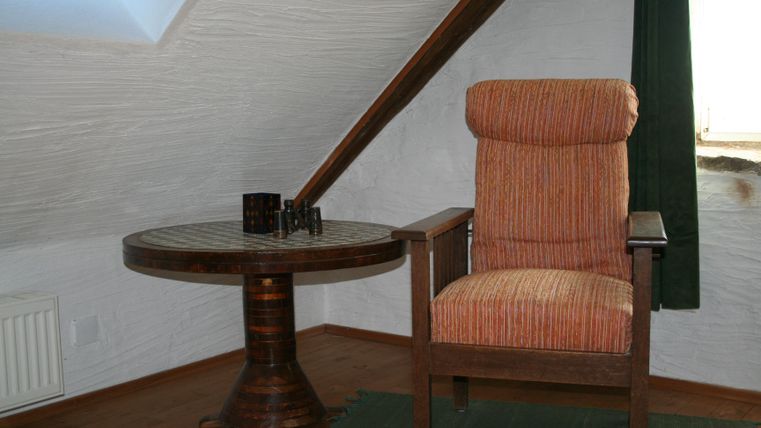 Een gezellige kamer op zolder met een houten stoel en een ronde tafel. De ramen laten natuurlijk licht binnen en er is een groene gordijn.
