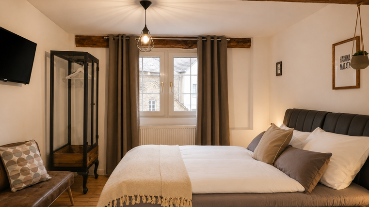 Een gezellige slaapkamer met een groot bed, elegante bedlinnen en stijlvolle decoratieve elementen. Een raam laat natuurlijk licht binnen en een decoratieve kapstok complementeert de ruimte.
