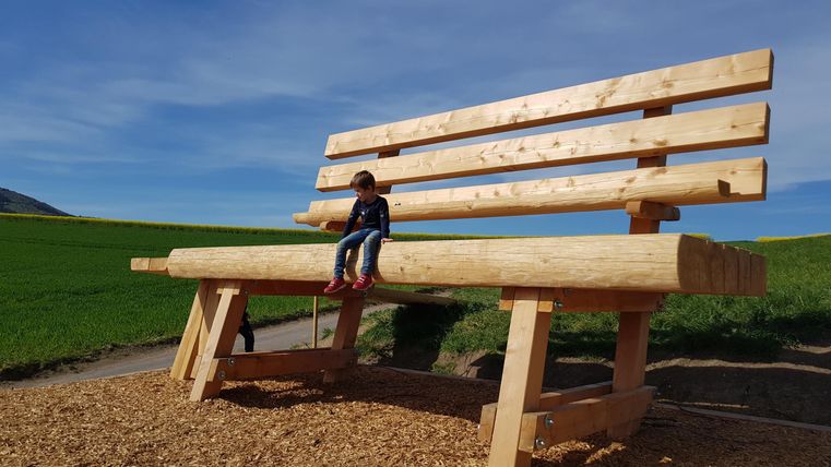 Een grote houten bank staat in een groene landschap. Een kind zit op de bank en geniet van het uitzicht.