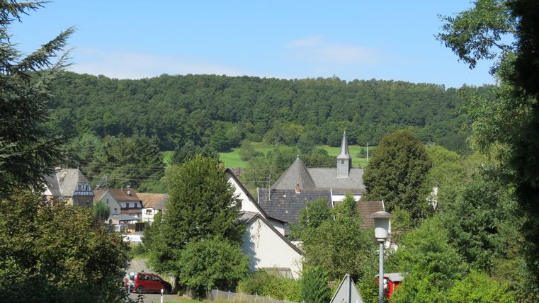 A picturesque village surrounded by lush greenery and gentle hills. In the background, a church with a tall tower can be seen.