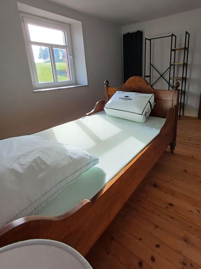 Une chambre simple avec un lit en bois et un grand coussin. La pièce a des murs clairs et un bureau de fenêtre qui permet de profiter de la lumière du jour.