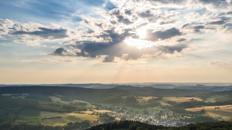 Landschapsgezicht van Neroth met beboste heuvels en een dorp in de vallei, onder een lucht met wolken en zonnestralen.
