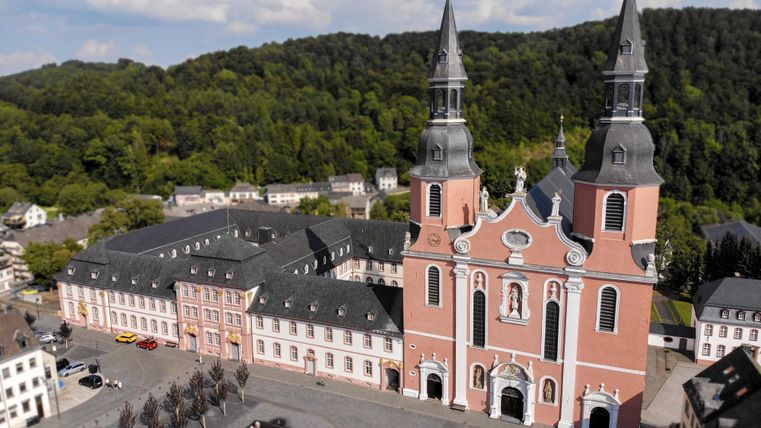Une église impressionnante avec deux tours et une façade colorée se dresse à côté d'un bâtiment historique. En arrière-plan, on aperçoit des collines boisées.