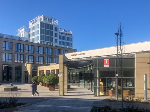 Ein modernes Gebäude mit großen Fenstern und einer klaren Architektur. Im Vordergrund ist eine Person zu sehen, die einen Gehweg entlanggeht.