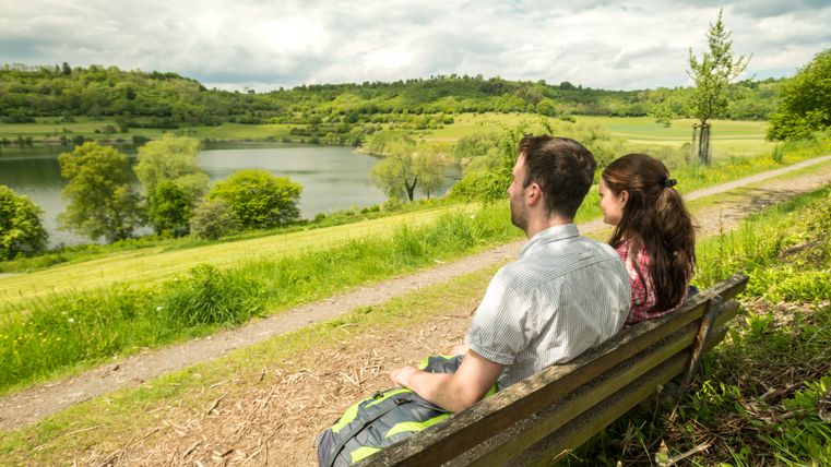 A couple sits on a bench and looks out over the Schalkenmehrener Maar, surrounded by green countryside and a cloudy sky.