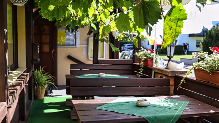 Ein gemütlicher Außenbereich mit Holztischen und grünen Tischdecken. Über den Tischen hängen üppige Blätter und Blumen in Töpfen.