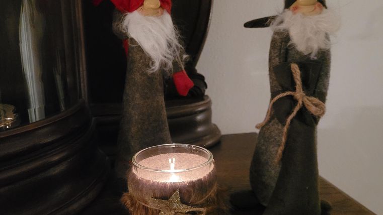 Zwei dekorative Figuren mit roten und grauen Hüten stehen neben einer Kerze auf einem Tisch. Die Kerze ist in einem rustikalen Halter platziert und verbreitet ein gemütliches Licht.