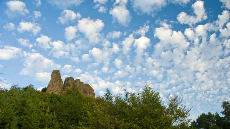 Een rotsachtige verhoging omgeven door bomen en een heldere blauwe lucht. Witte, pluizige wolken trekken voorbij aan de lucht.