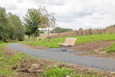 A wooden bench by the side of the path, surrounded by trees, a field and houses