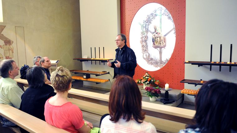 Bright interior of a modern church. People are sitting on one of the wooden benches, a priest can be seen in the foreground.