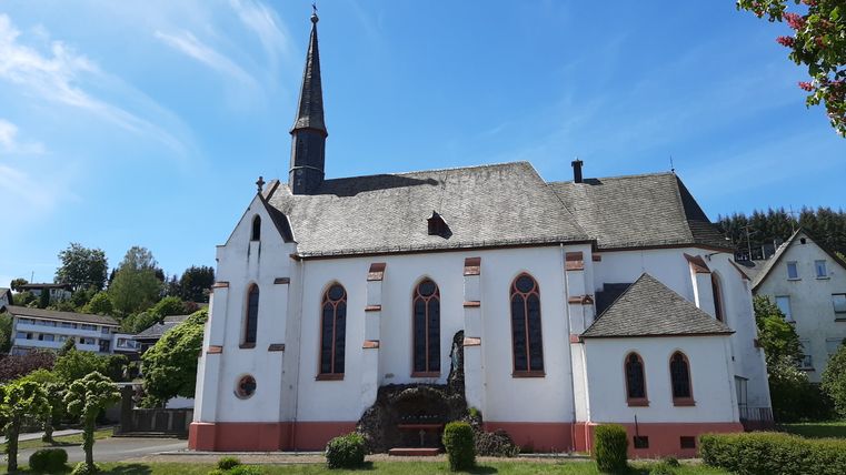 Witte kerk met spitse toren en rode ramen in Deudesfeld, omringd door bomen en blauwe lucht.