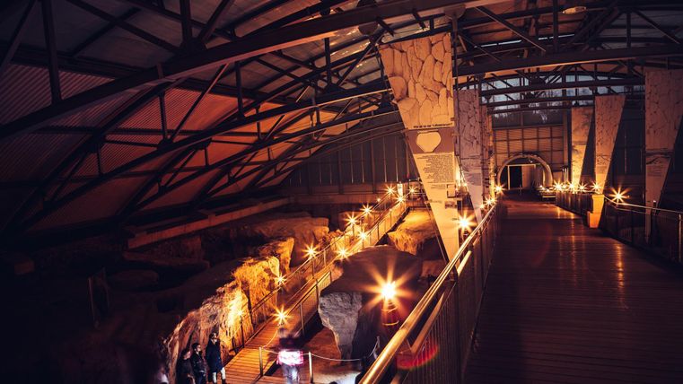 Eine moderne Ausstellungshalle mit einem Holzsteg und sanfter Beleuchtung. Im Hintergrund sind archäologische Funde und Besucher zu sehen.