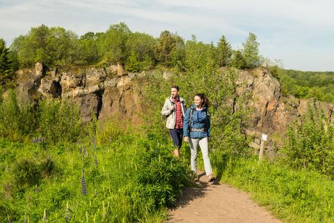 Two hikers in the volcanic garden Steffeln in front of the large rock wall of the volcano Steffelberg surrounded by green bushes and trees.