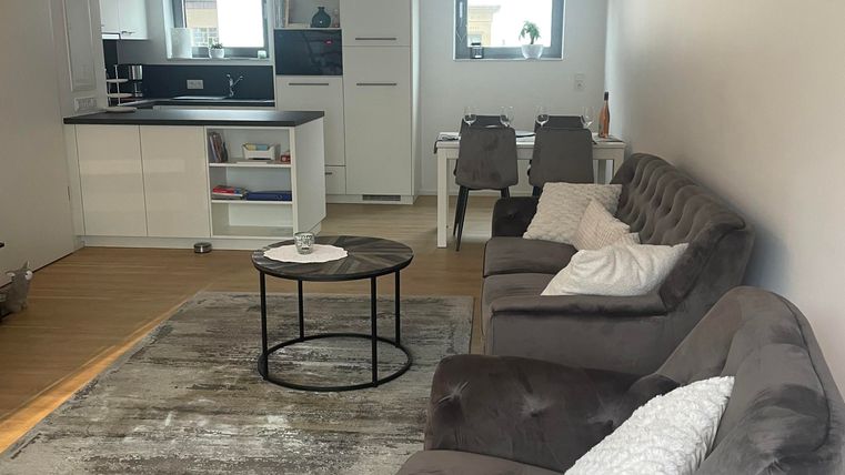 Ein modernes Wohnzimmer mit grauen Sofas und einem runden Tisch. Die offene Küche ist gut ausgestattet und der Raum ist hell und einladend.