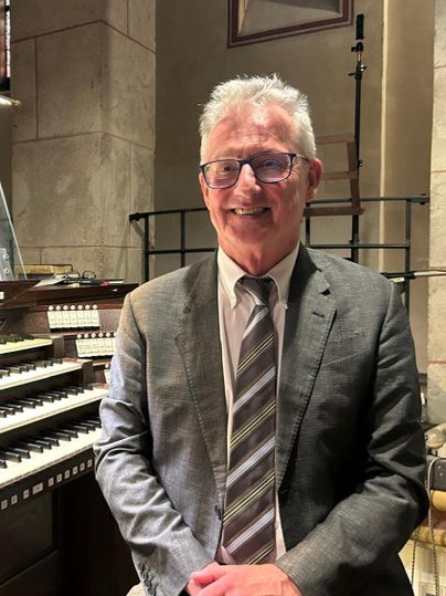 Ein lächelnder Mann in einem Anzug sitzt vor einer Orgel. Die Umgebung wirkt musikalisch und feierlich.