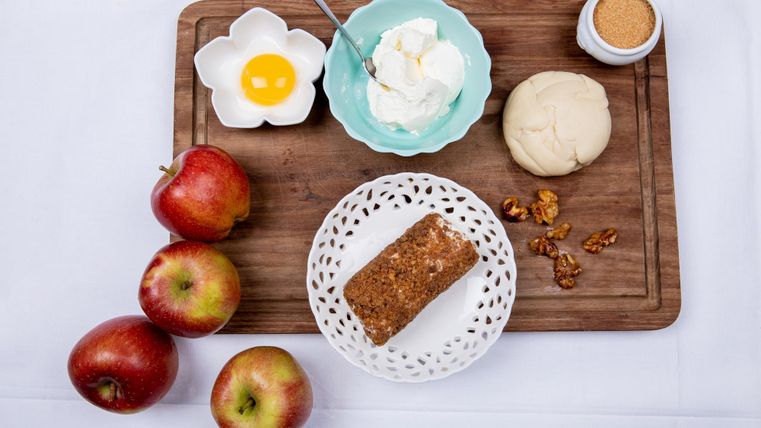 Een houten plank met verse appels, een stuk taart en een kom met room. Daarnaast zijn een rauw ei, een stuk deeg en een glas suiker te zien.