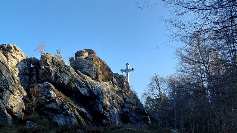 Un beau rocher avec une croix, entouré d'arbres et d'un ciel dégagé. Le chemin traverse la nature.