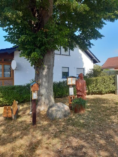 Rastplatz mit Baum, Holzfigur und Sitzbank vor einem Haus.
