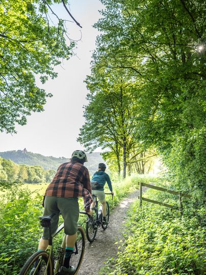 Two cyclists on gravel bikes ride along a narrow path next to a dense deciduous forest and wide meadows.