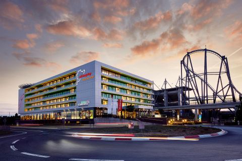 Een modern hotel met gekleurde ramen staat voor een opwindend pretpark. De lucht is in zachte avondkleuren gehouden.