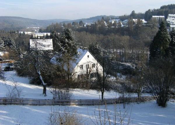 A snowy landscape with a white house and snow-covered trees. The tranquil setting is surrounded by gentle hills.