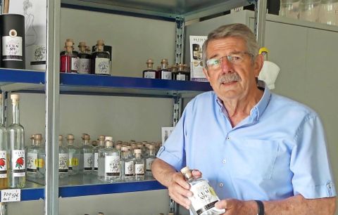 Een oudere man staat voor een rek met verschillende flessen. Hij houdt een fles vasthanden en kijkt vriendelijk in de camera.