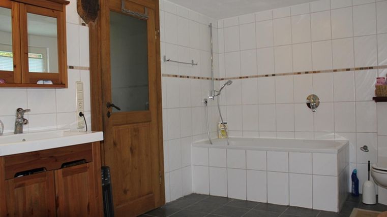 Een ruime badkamer met wit betegelde muren en een houten deur. Er is een bad, een douche en een dubbel wastafel.