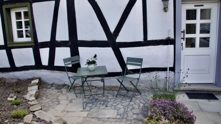 Ein gemütlicher Außenbereich mit einem kleinen Tisch und zwei Stühlen. Die Wand ist im Fachwerkstil gestaltet und das Blumenbeet sorgt für eine freundliche Atmosphäre.