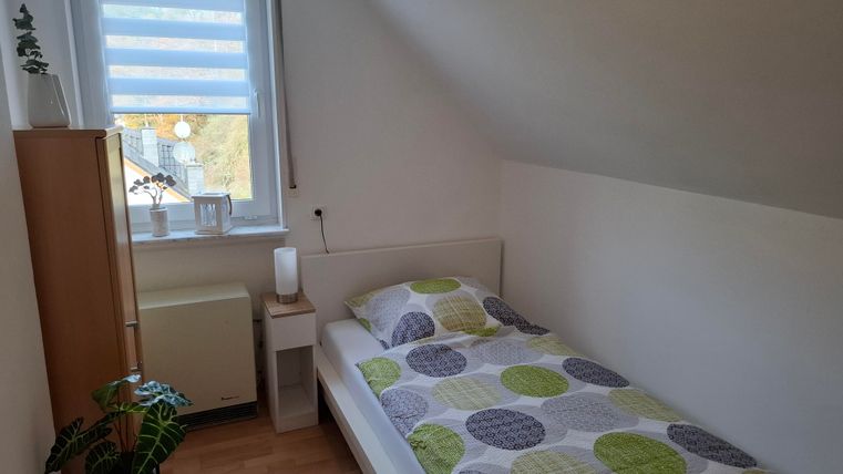 Een kleine, lichte slaapkamer met een eenpersoonsbed en kleurrijke kussens. Naast het bed staat een nachttafeltje en een rek, en een raam zorgt voor daglicht.