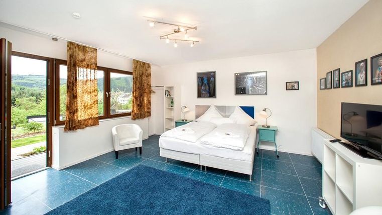 Bright and modern bedroom with a large double bed and a blue carpet. Windows offer a view of nature and the decor is minimalistic.