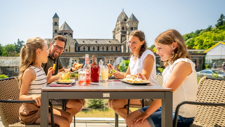 Familie mit zwei Kindern sitzt bei sonnigem Wetter auf der Terrasse einer Gaststätte und genießt ein gemeinsames Essen. Im Hintergrund die eindrucksvolle romanische Basilika Maria Laach mit ihren markanten Türmen. Auf dem Tisch stehen bunte Getränke und verschiedene Speisen. Die Stimmung ist fröhlich und entspannt.