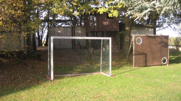 A soccer goal on a green meadow, surrounded by trees. In the background, a playhouse is visible.