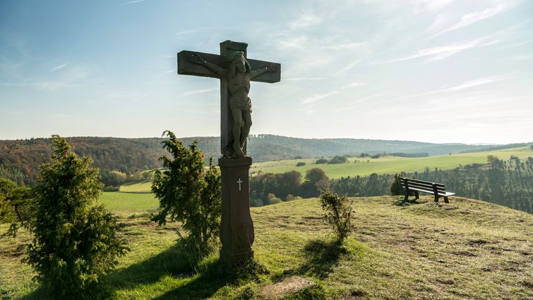 Kreuz auf einem Hügel mit Bank und weiter Landschaft im Hintergrund.