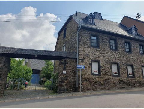 A historical stone building with a distinctive bay window. In the foreground, trees and an entrance area can be seen.