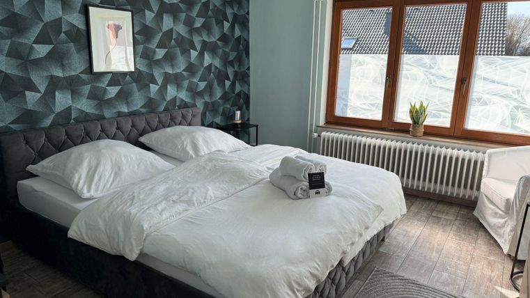 A modern bedroom with a large double bed and a geometric wall design. The room has large windows that let in natural light.