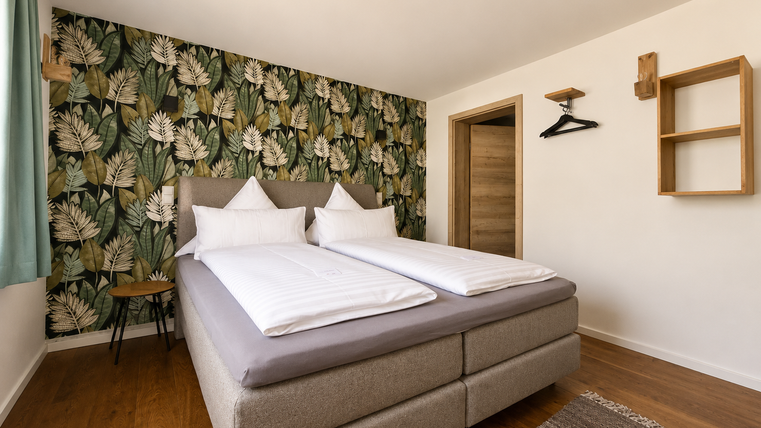 Een moderne slaapkamer met een tweepersoonsbed en een stijlvolle behang met plantmotief. De kamer is licht en vriendelijk ingericht met een houten vloer en minimalistisch meubilair.